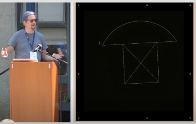 Adam delivers a talk, gesturing while at a podium in front of a screen showing the design of a rivet.